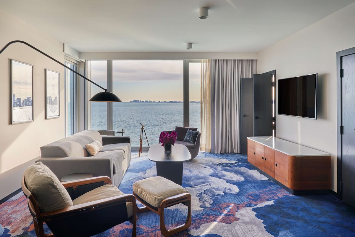 A modern hotel room with a large window offering a city view, a flat-screen TV, a sofa, a coffee table, and a wooden cabinet.