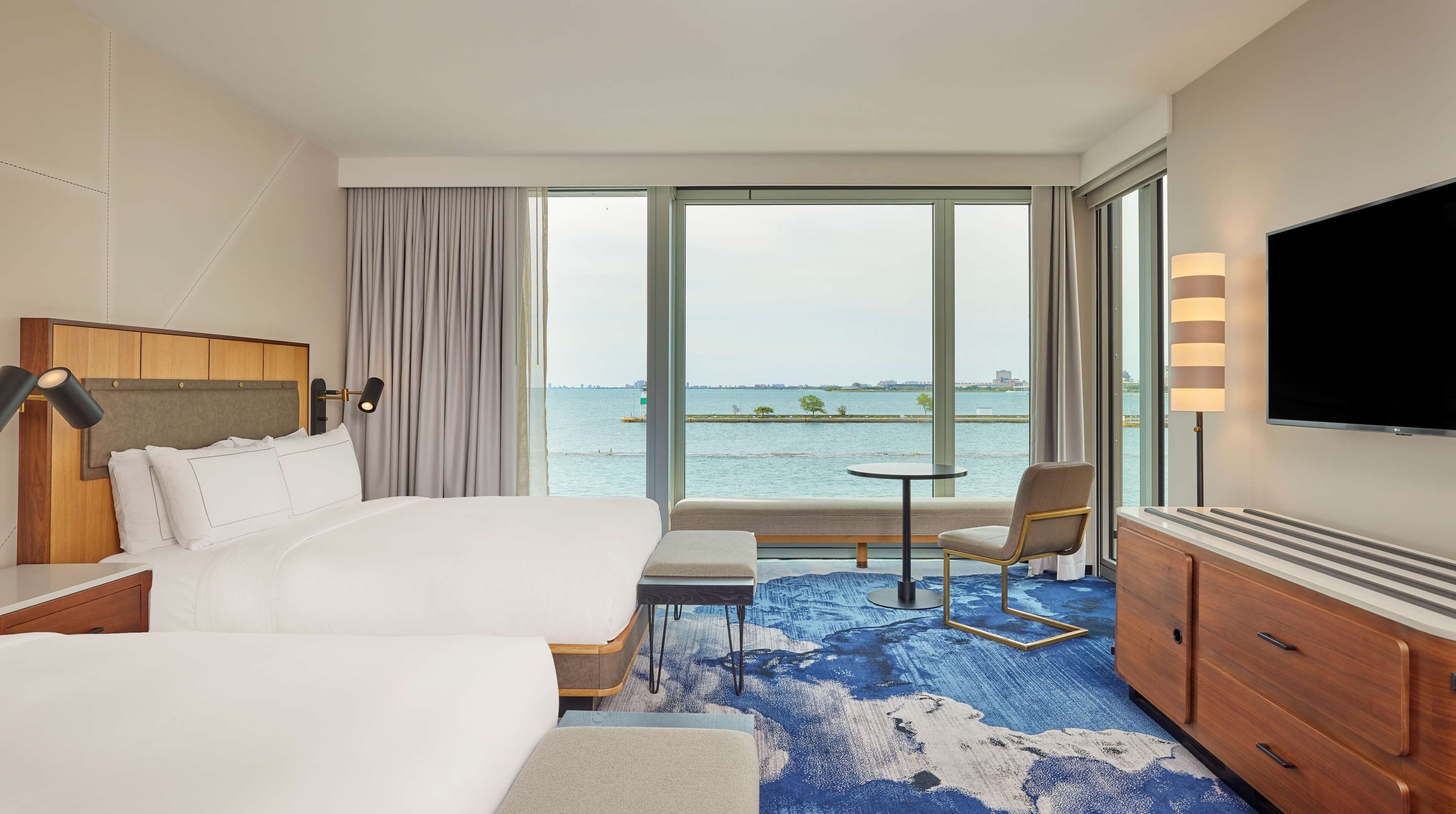 A hotel room with a large bed, a desk, a chair, and a TV. There is a view of the water and a cityscape through the windows.