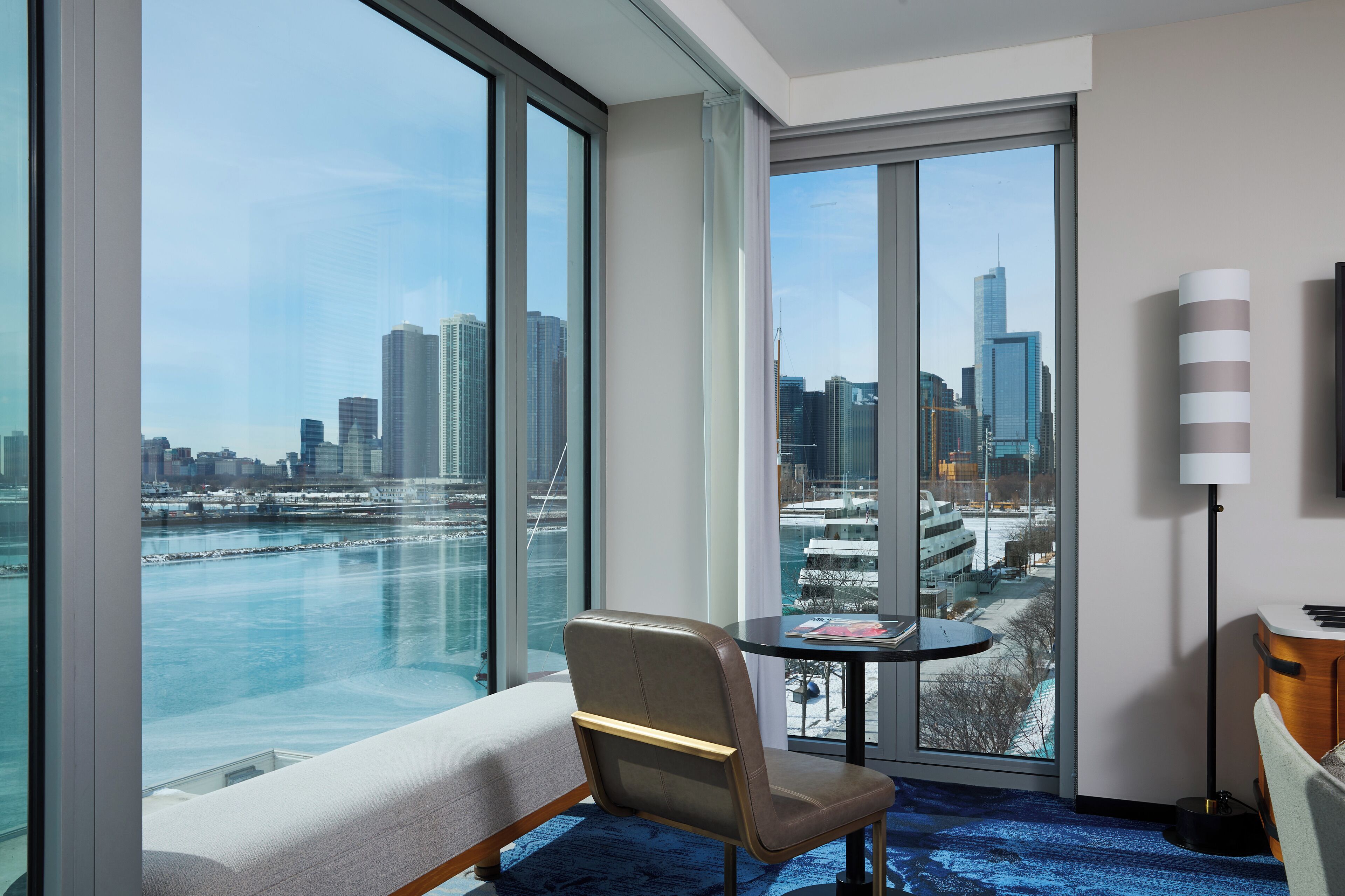 Quarto de hotel com uma janela grande que oferece vista da cidade, uma mesa redonda pequena, uma cadeira e um abajur.