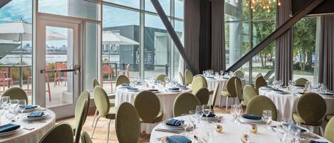 A restaurant with round tables set for dining, large windows, and a modern chandelier.