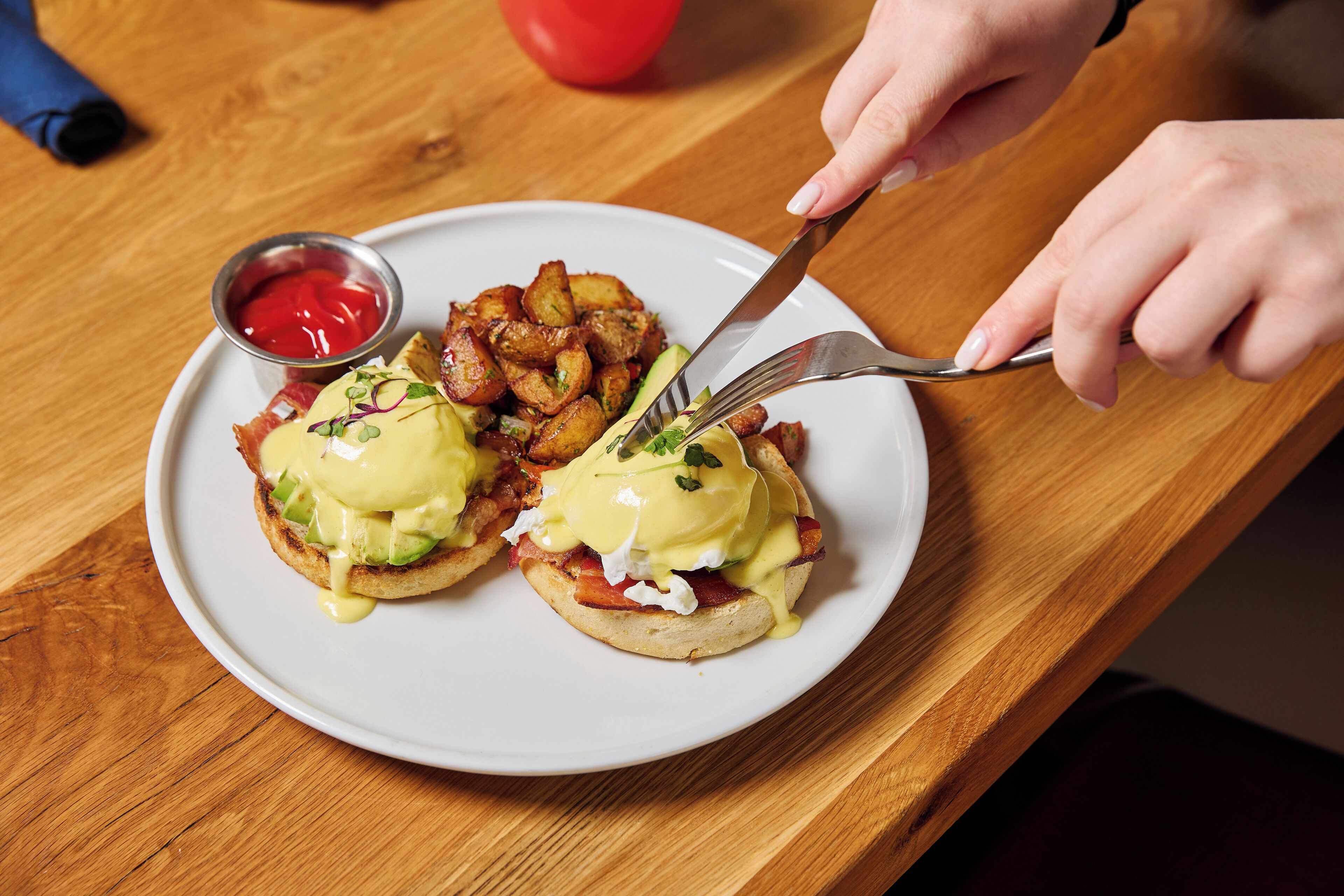 Un plato con dos sándwiches huevos benedictinos, patatas ralladas y un recipiente pequeño de kétchup.