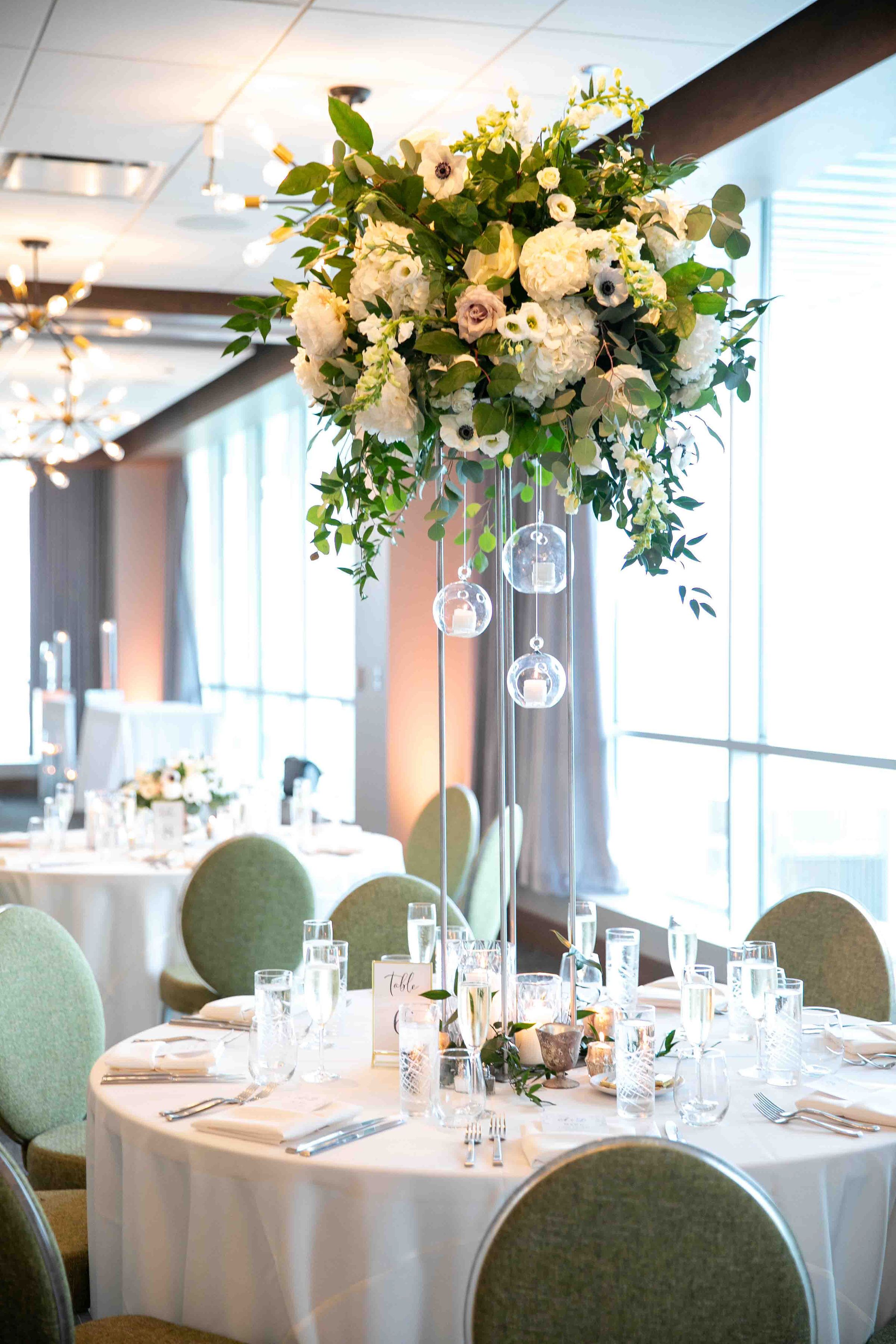 Een balzaal met ronde tafels gedekt voor een formeel evenement, met bloemstukken als decoratie en hangende kristallen bollen.