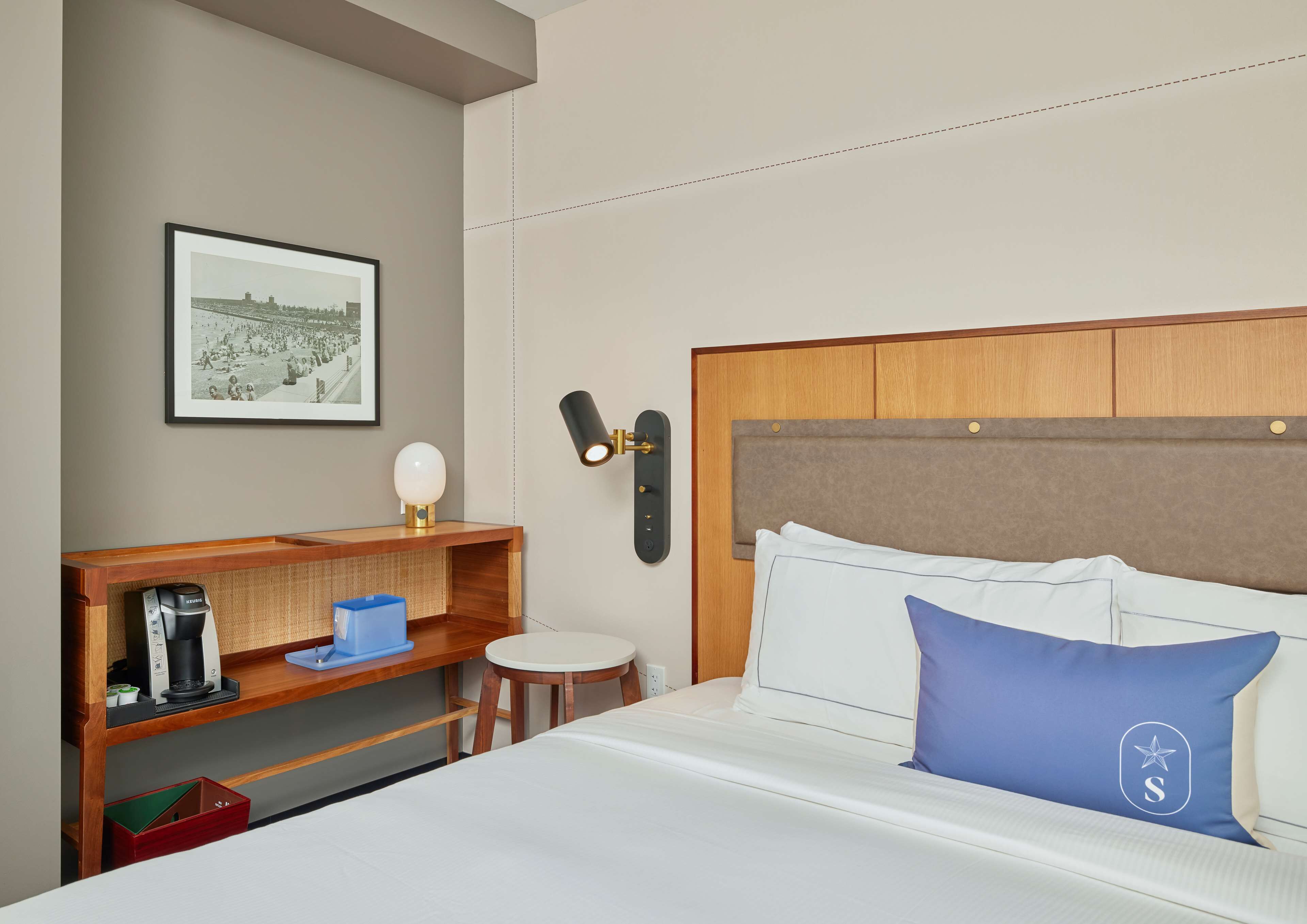 A hotel room with a bed, a bedside table with a lamp, a framed picture on the wall, and a small stool.