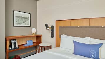 A hotel room with a bed, a bedside table with a lamp, a framed picture on the wall, and a small stool.