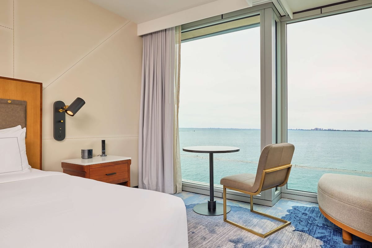 A hotel room with a bed, a bedside table with a lamp, a chair by the window, and a view of the sea.