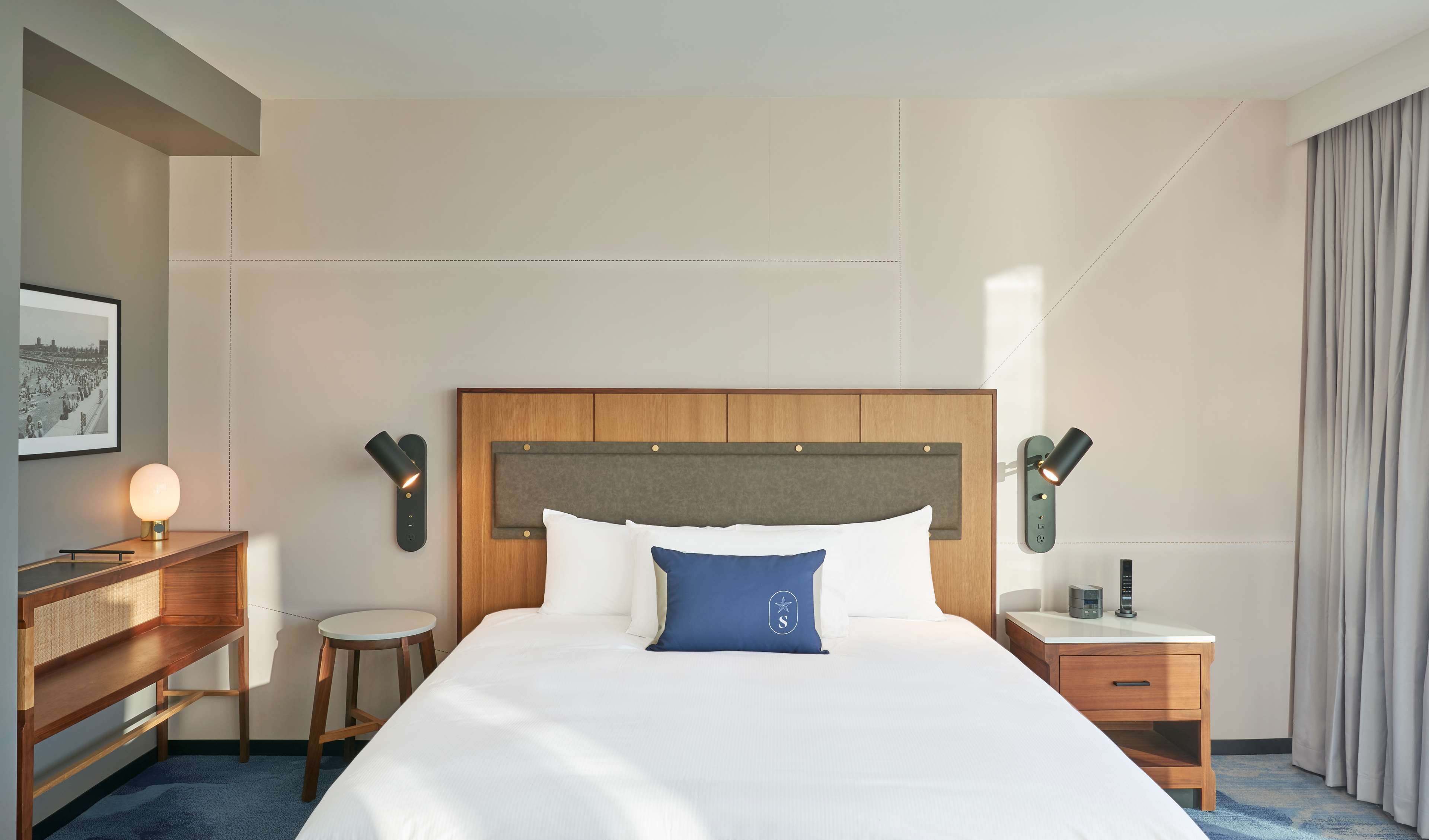 Habitación de hotel con una cama grande, cabecera de madera, mesitas de noche, una lámpara, un reloj y un cuadro en la pared.