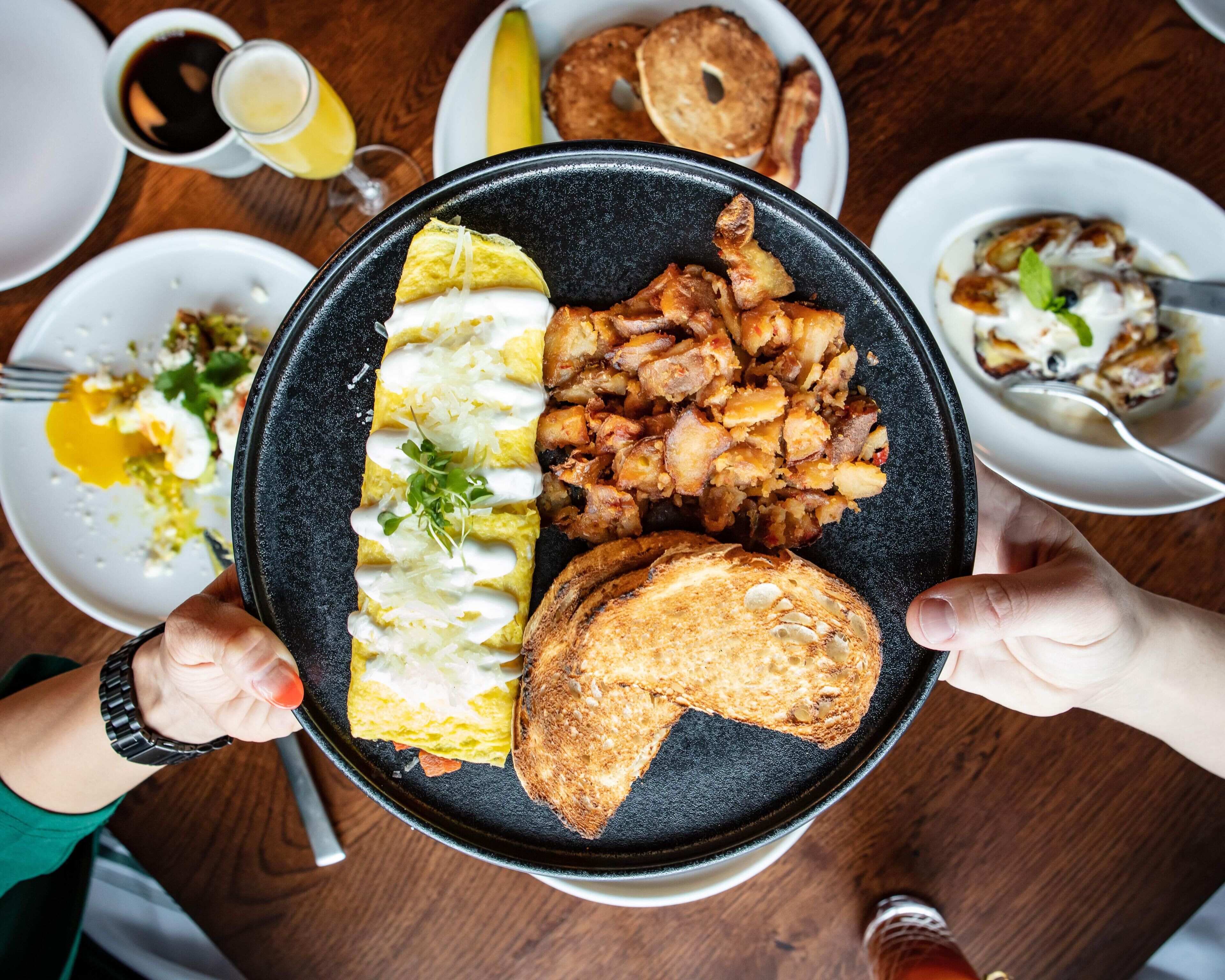 A person holding a black plate with a breakfast meal consisting of eggs, hash browns, toast, and a side salad.