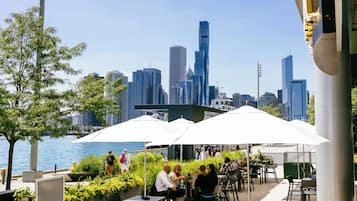 An outdoor seating area with umbrellas, tables, and chairs, overlooking a city skyline and a body of water.