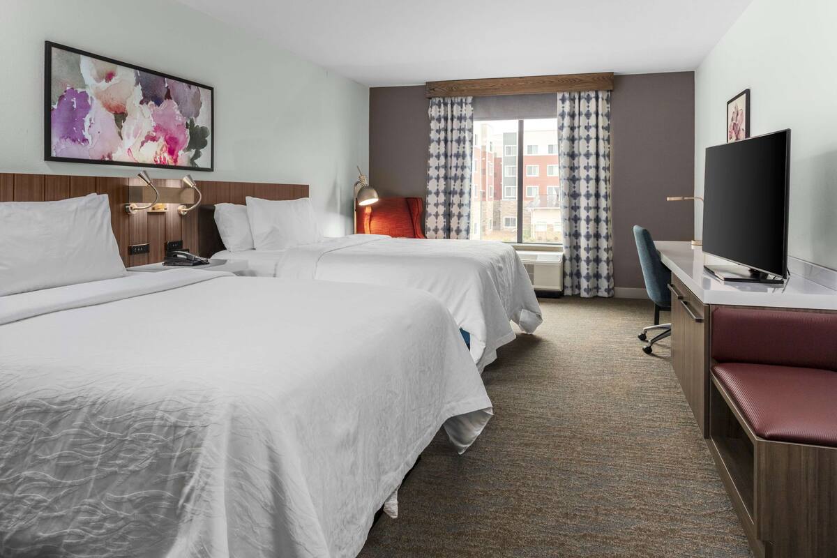 A hotel room with two beds, a desk, a TV, and a window with curtains.