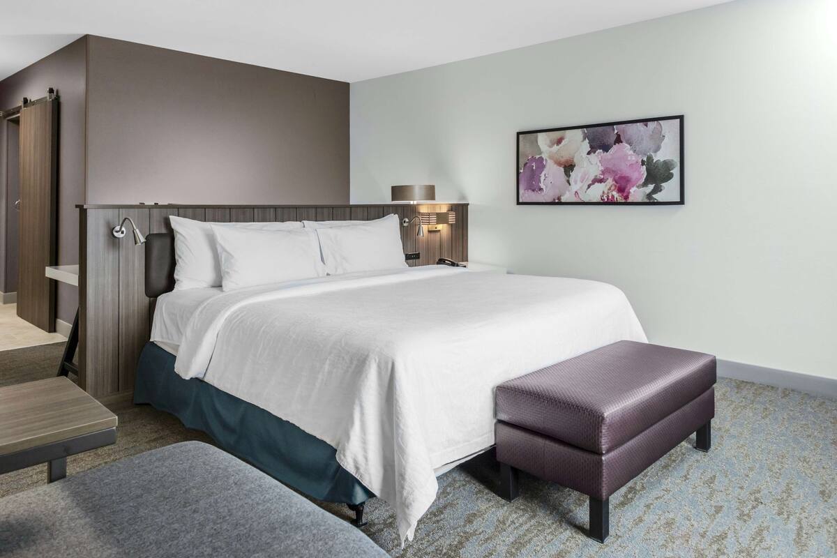 A modern hotel room with a large bed, a bench, a nightstand, and a wall-mounted lamp.