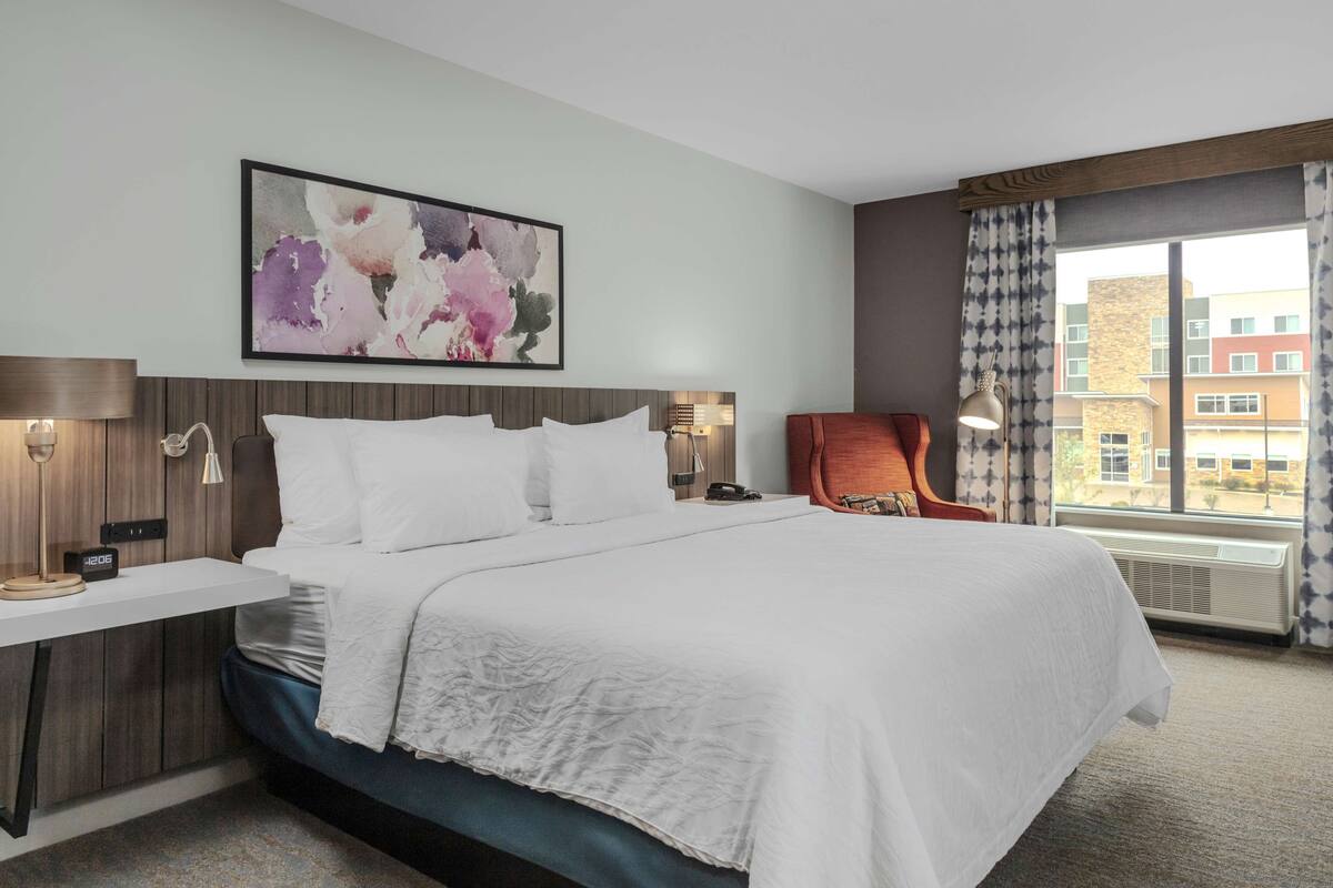 A hotel room with a large bed, a bedside table with a lamp, a chair, and a window with curtains.