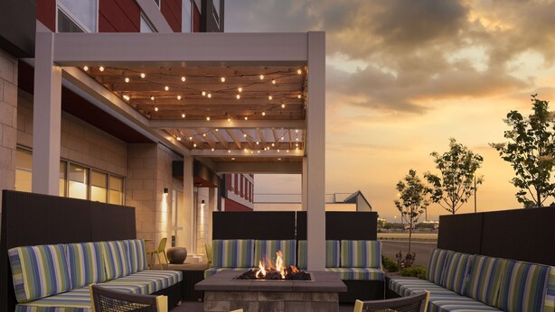 An outdoor seating area with a fire feature, cushioned seating, and string lights. - Home2 Suites by Hilton Fishers Indianapolis Northeast, IN (Fishers)