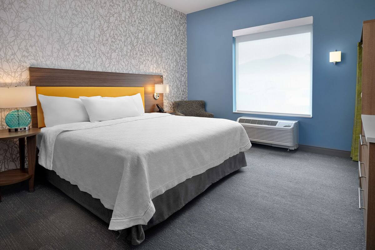 A hotel room with a bed, a nightstand, a lamp, a wall-mounted air conditioner, and a window with a view.