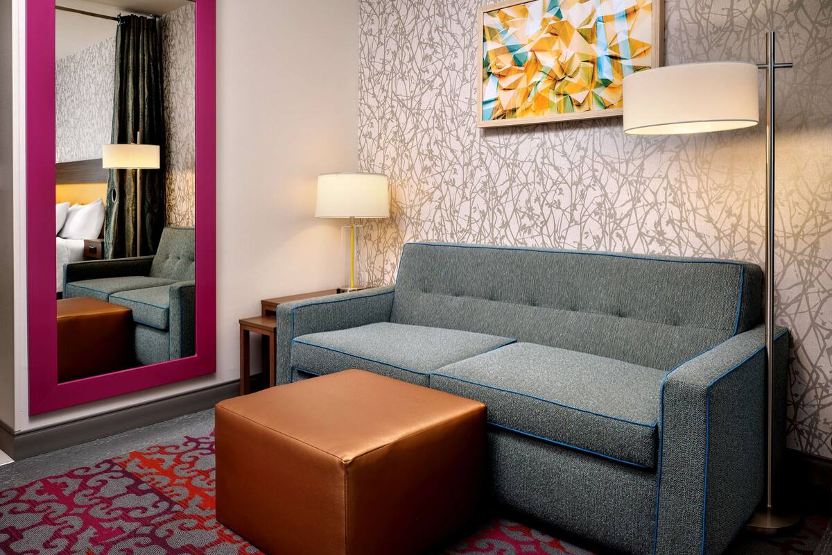 A modern hotel room with a grey sofa, a brown ottoman, a large mirror, a floor lamp, and a wall art piece.