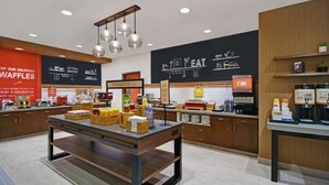 A modern cafe with a central wooden table, various food items, and a blackboard with menu options. - Hampton Inn Las Vegas Strip South, NV (Las Vegas)