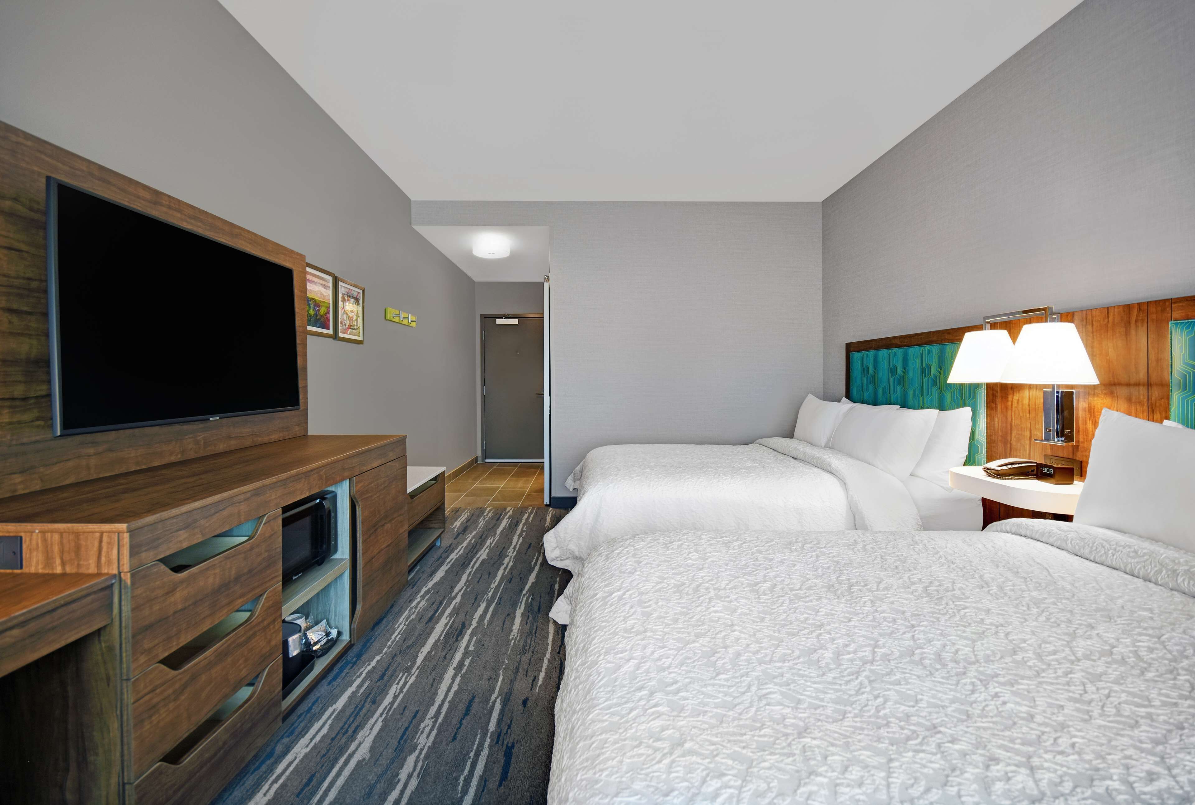 A hotel room with a flat-screen TV, a wooden entertainment unit, two beds with white bedding, a lamp, and a carpeted floor.