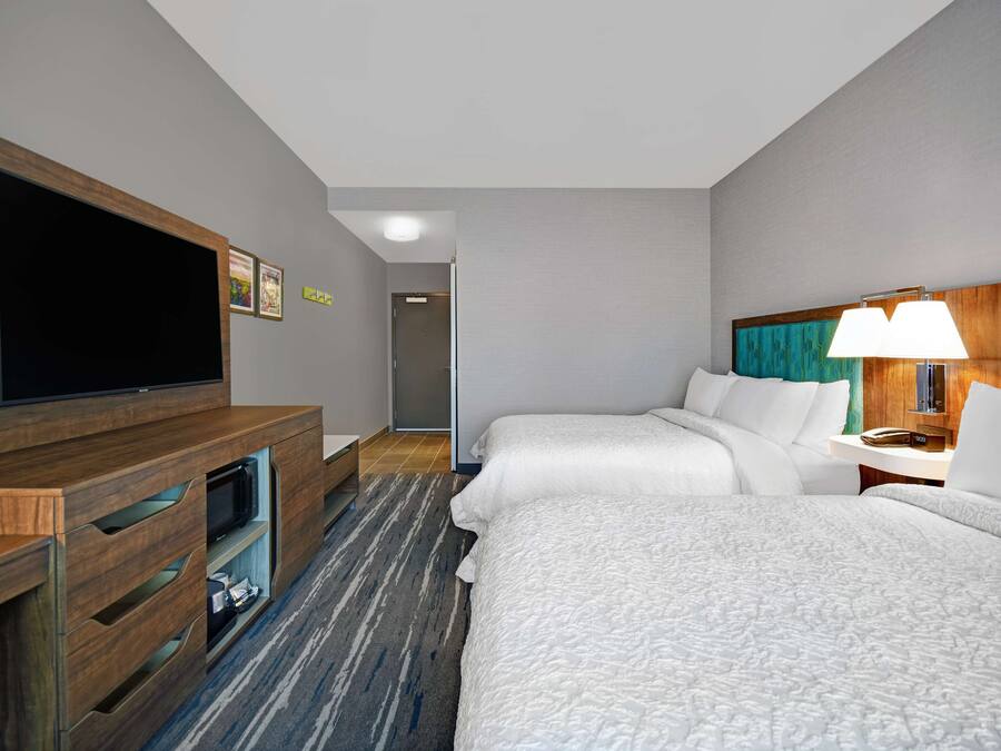 Habitación de hotel con televisor de pantalla plana, mueble de madera para entretenimiento, dos camas con ropa blanca, lámpara y piso alfombrado.