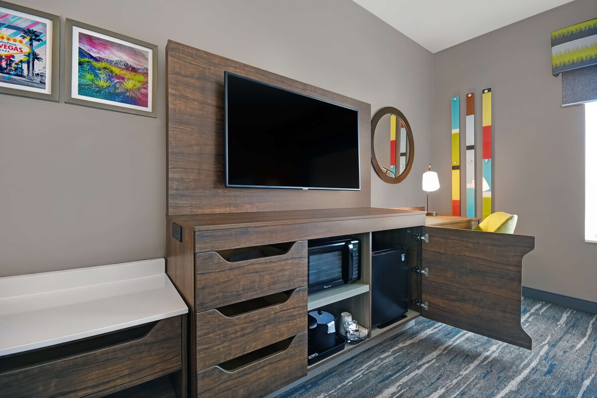 A modern hotel room with a flat-screen TV mounted on a wooden cabinet, a microwave, and a small refrigerator. There are two framed pictures on the wall and a colorful abstract painting.