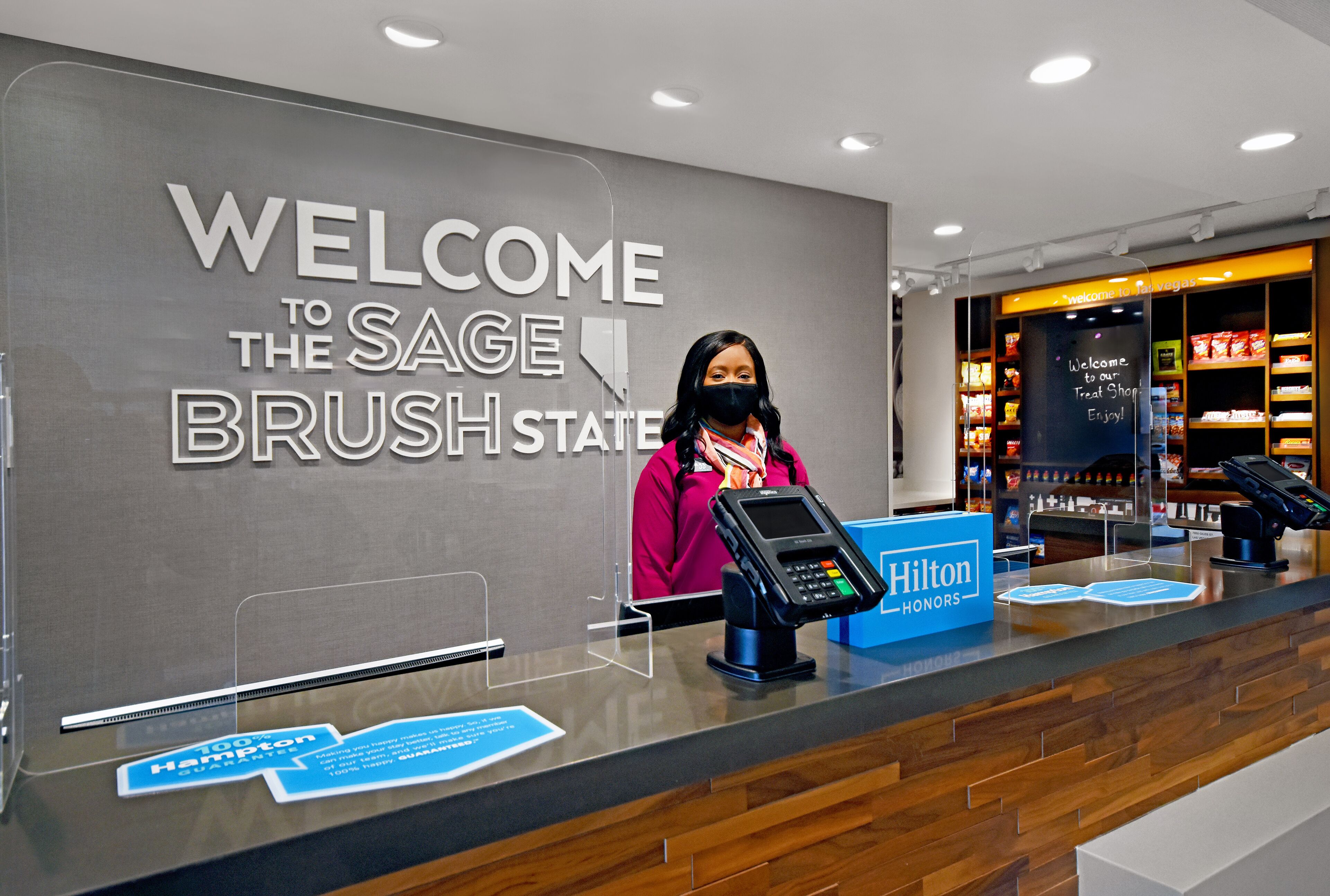 Un comptoir d’accueil d’hôtel avec une grande enseigne indiquant « Bienvenue », un terminal de paiement numérique et un panneau indiquant que l’hôtel fait partie du programme Hilton Honors.