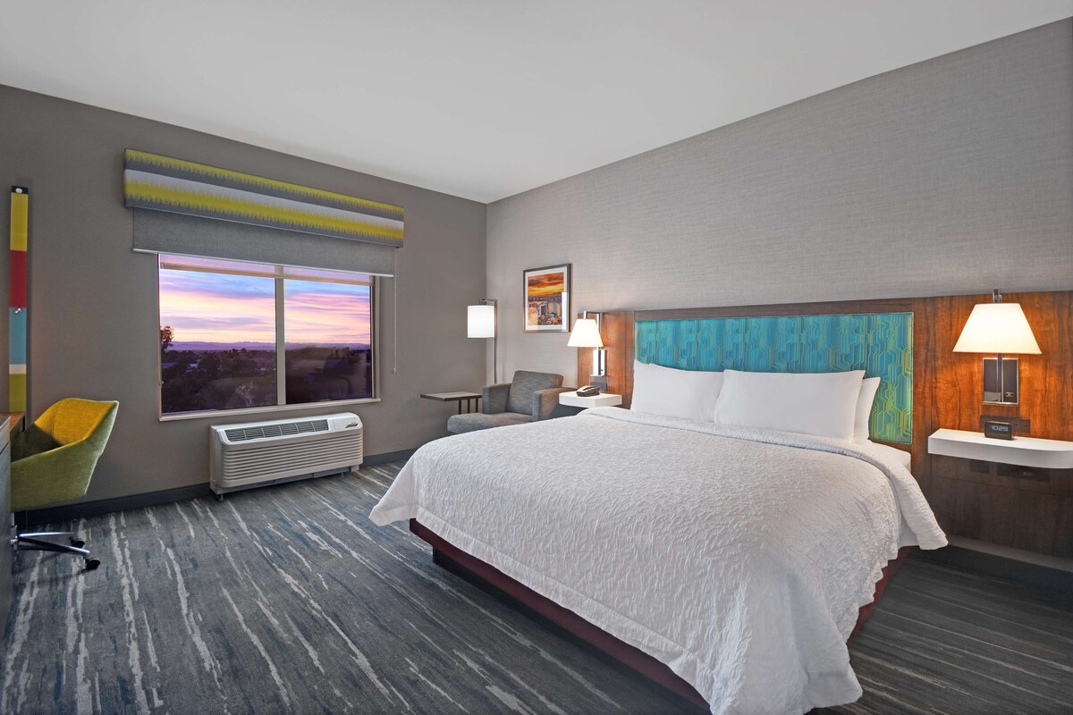 A modern hotel room with a large bed, a view of the sunset, a desk with a chair, and a small table with a lamp.