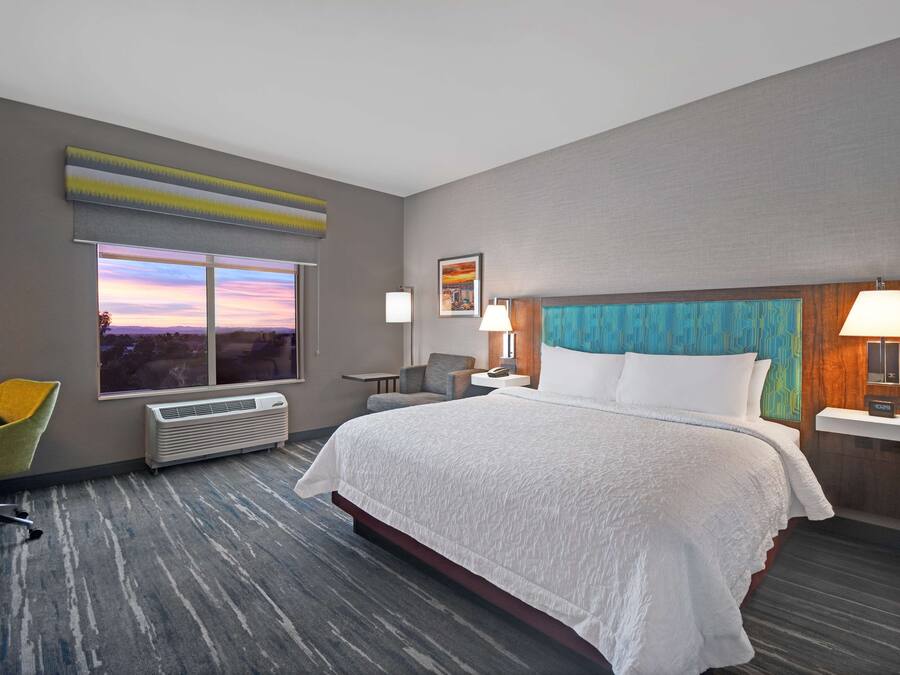 Ein modernes Hotelzimmer mit einem großen Bett, Blick auf den Sonnenuntergang, einem Schreibtisch mit Stuhl und einem kleinen Tisch mit Lampe.