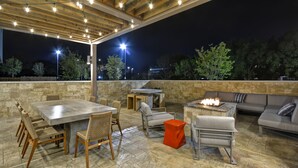 En udendørs terrasse med klinkegulv, træpergola, lyskæder, en bålplads og siddepladser.