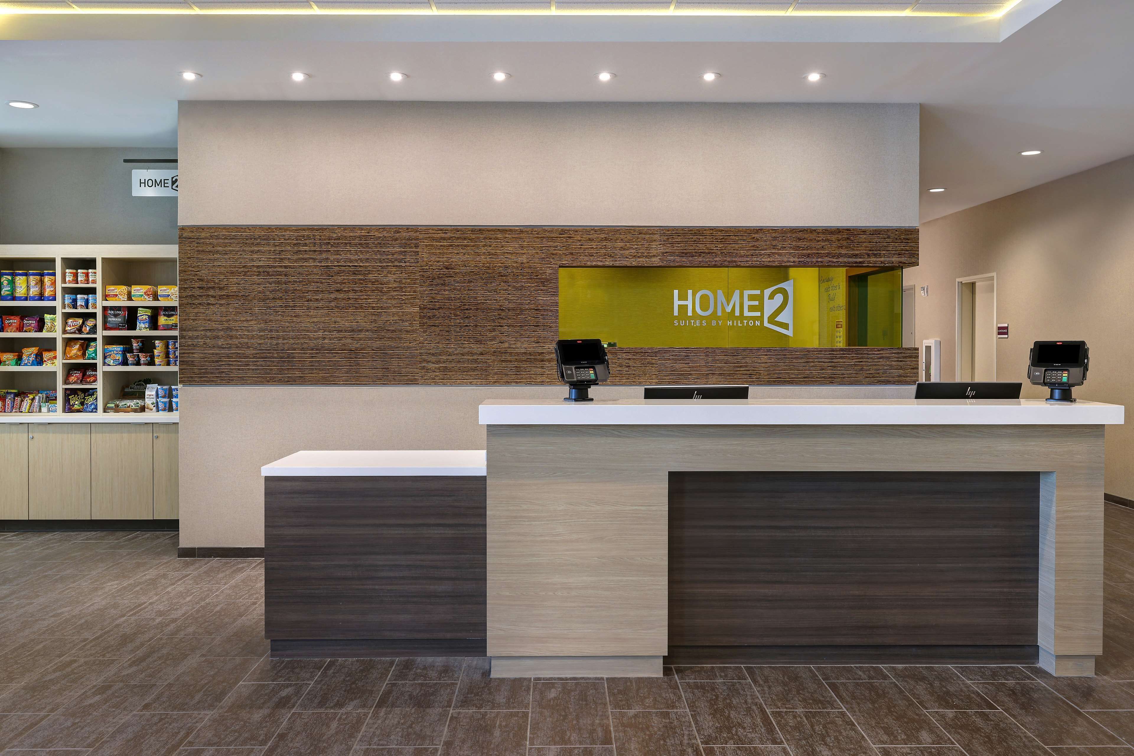 Un comptoir d’accueil d’hôtel avec une enseigne « HOME2 », un comptoir et une enseigne « HOME » au-dessus d’une étagère contenant des en-cas.