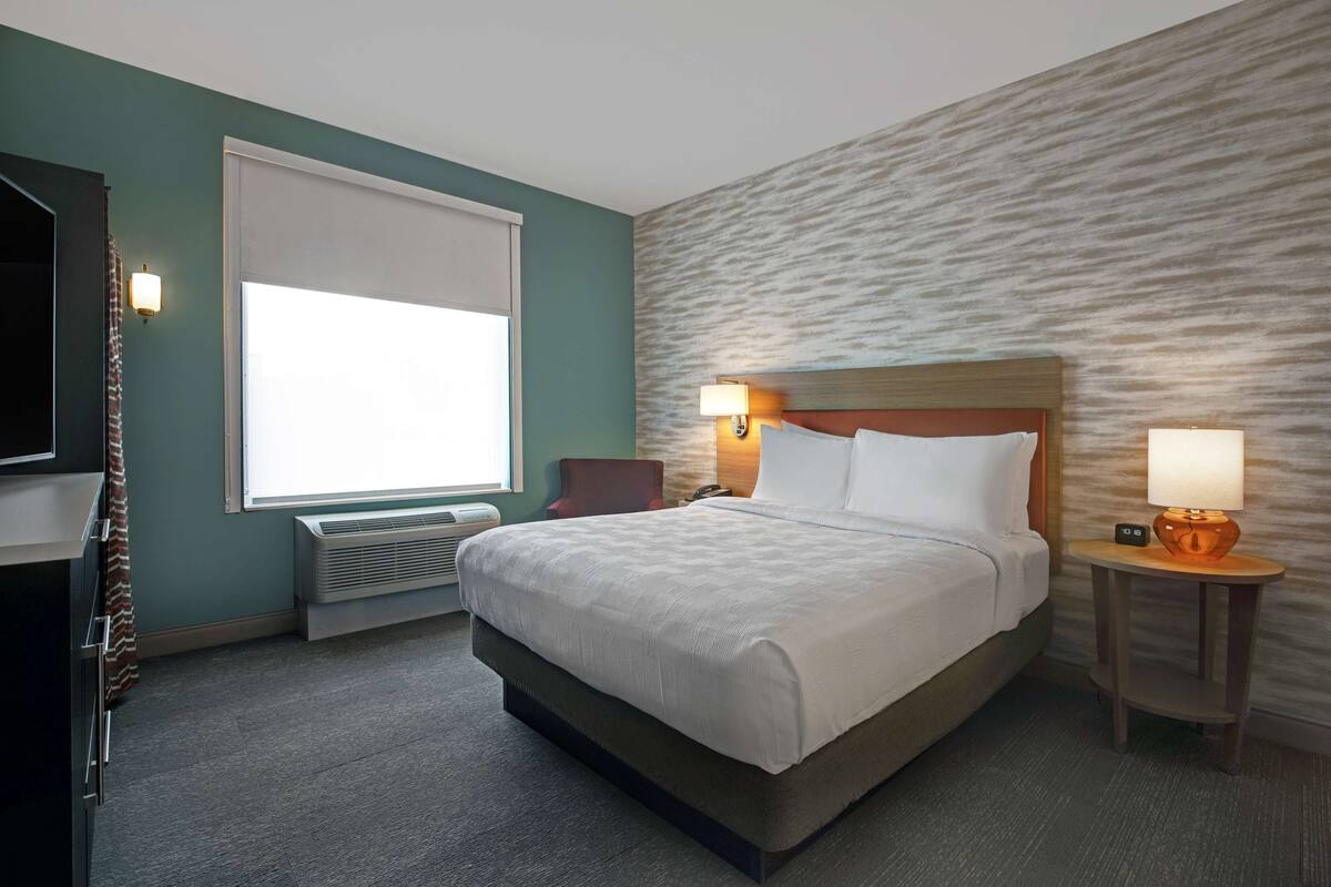 Een hotelkamer met een groot bed, twee nachtkastjes met lampen, een flatscreen televisie en een raam met zonwering.