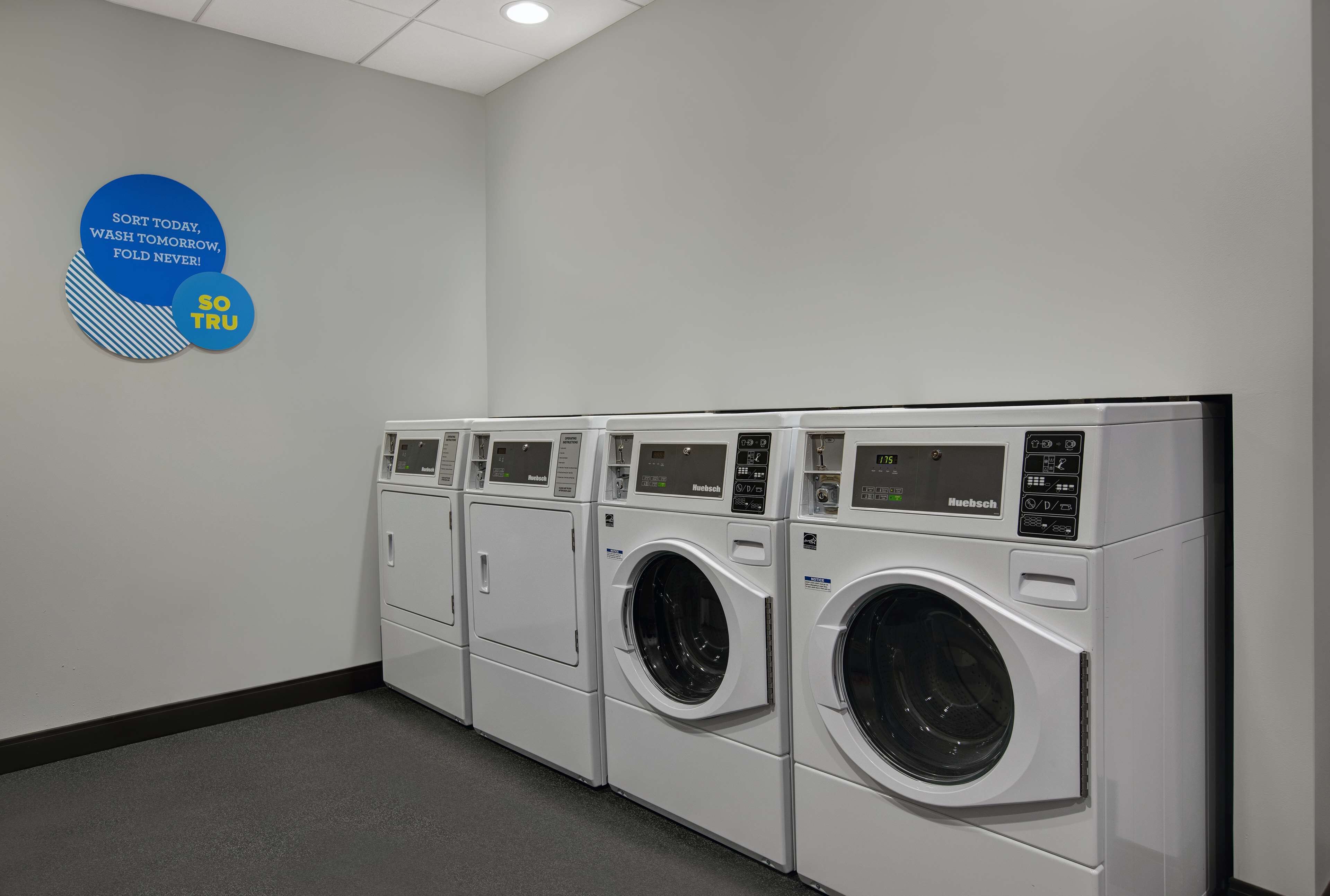 A laundry facility with multiple front-loading washing machines, a wall-mounted sign with motivational text, and a grey carpeted floor.