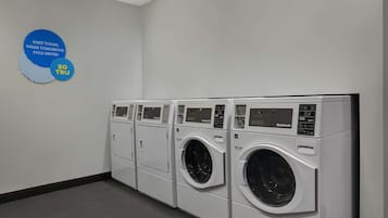 A laundry facility with multiple front-loading washing machines, a wall-mounted sign with motivational text, and a grey carpeted floor.