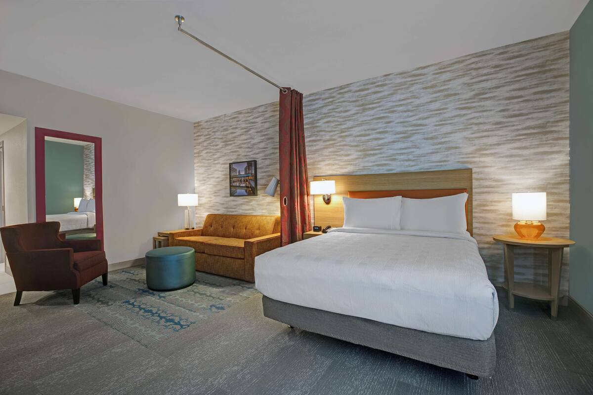 Een hotelkamer met een groot bed, een houten hoofdbord, twee nachtkastjes met lampen, een bruine fauteuil, een blauwe poef en een rood gordijn.