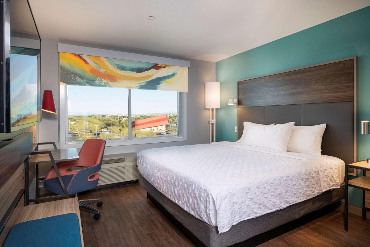 A modern hotel room with a large bed, a desk, and a chair. There is a window with a view of the city.