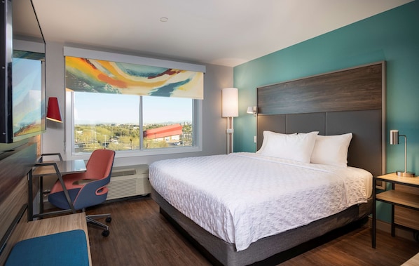 Habitación de hotel moderna con una cama grande, un escritorio con silla, una ventana con vistas y un cuadro abstracto colorido.