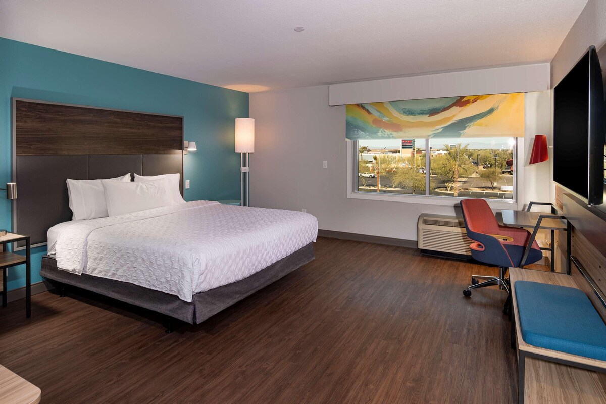 A modern hotel room with a large bed, a desk, a chair, and a window with a view of a parking lot.