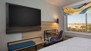 A hotel room with a large flat-screen TV mounted on the wall, a bed with a white mattress, a blue bench, a red and blue chair, and a window with a city view.