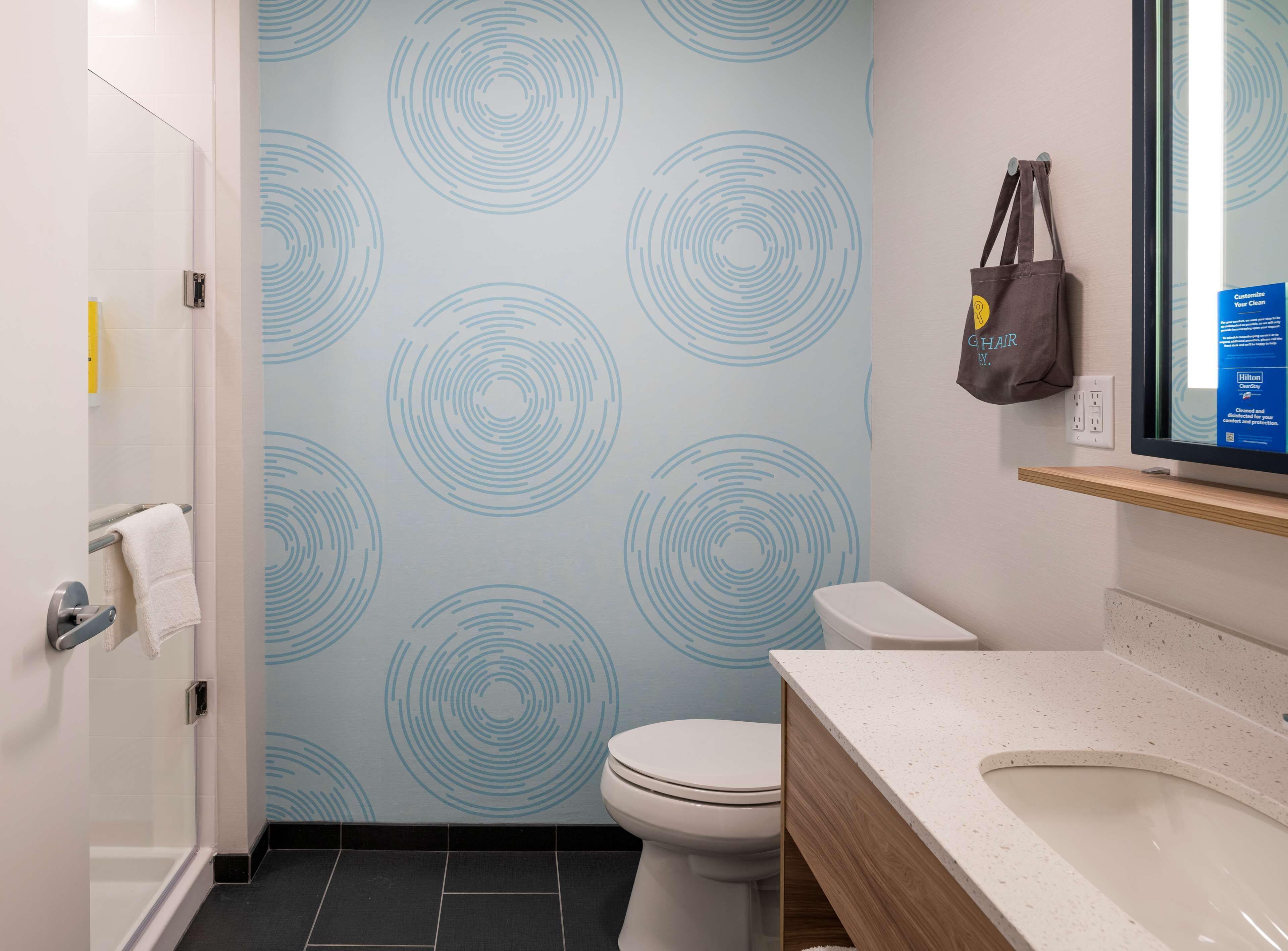 Une salle de bain avec des toilettes, un lavabo et une douche. On y trouve un porte-serviettes sur lequel est accrochée une serviette. Les murs sont décorés d’un motif circulaire.