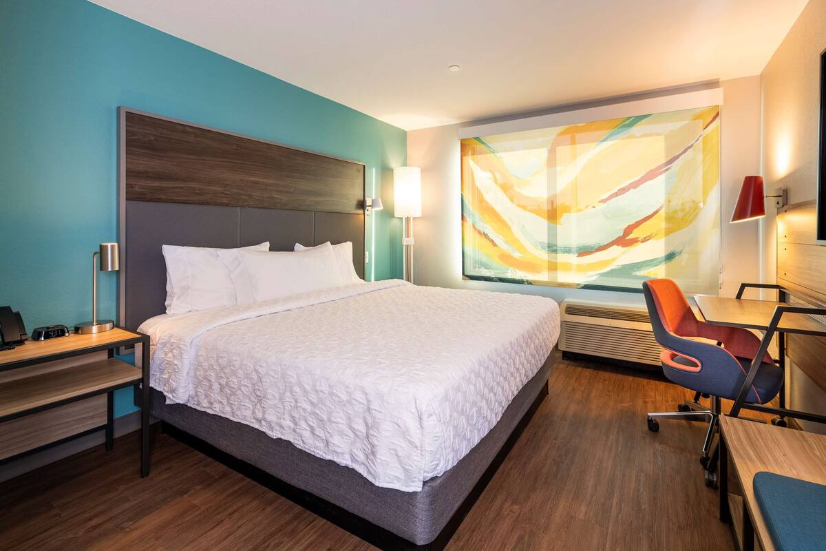 Ein Hotelzimmer mit einem großen Bett, einem Schreibtisch mit Stuhl, einem Fernseher und einem großen, abstrakten Gemälde an der Wand.