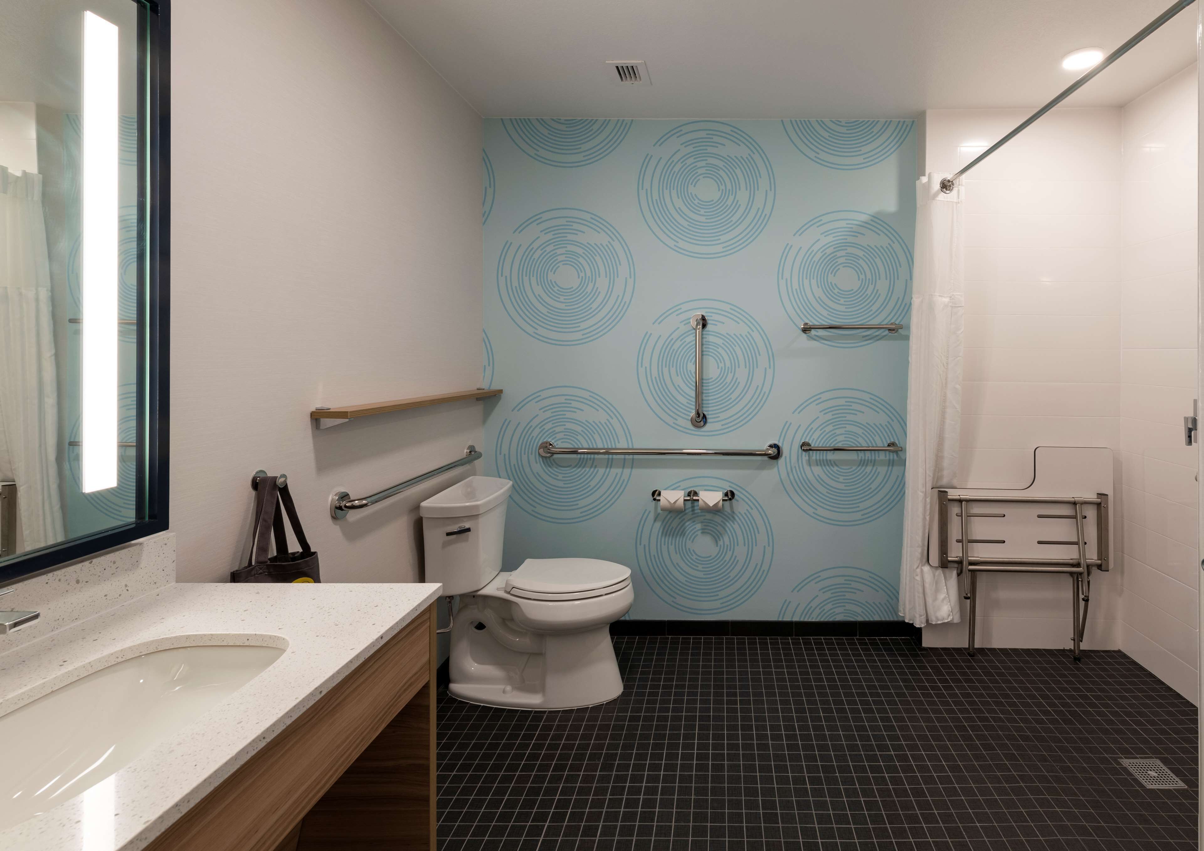Un baño moderno con inodoro blanco, lavamanos con encimera moteada y espejo. Hay pasamanos en las paredes y una zona de ducha con cortina.