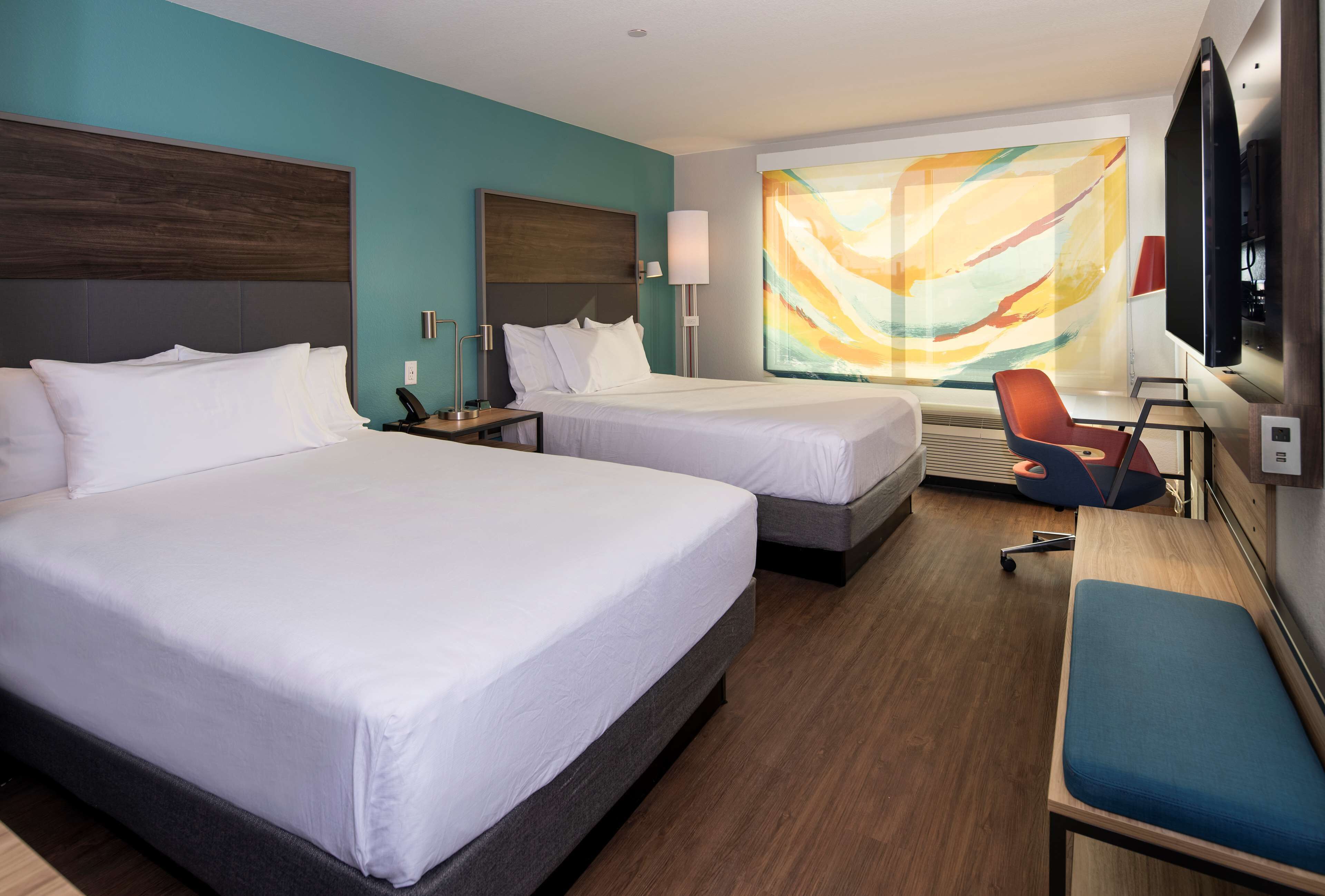 Quarto de hotel com duas camas, uma escrivaninha, uma cadeira, uma televisão e uma janela grande com vista colorida.