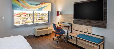 A modern hotel room with a large bed, a desk with a chair, a TV mounted on the wall, and a window with a view of the city.