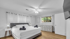 Une chambre à coucher comprenant un lit, des tables de chevet, un ventilateur de plafond, une fenêtre avec des rideaux et une commode.