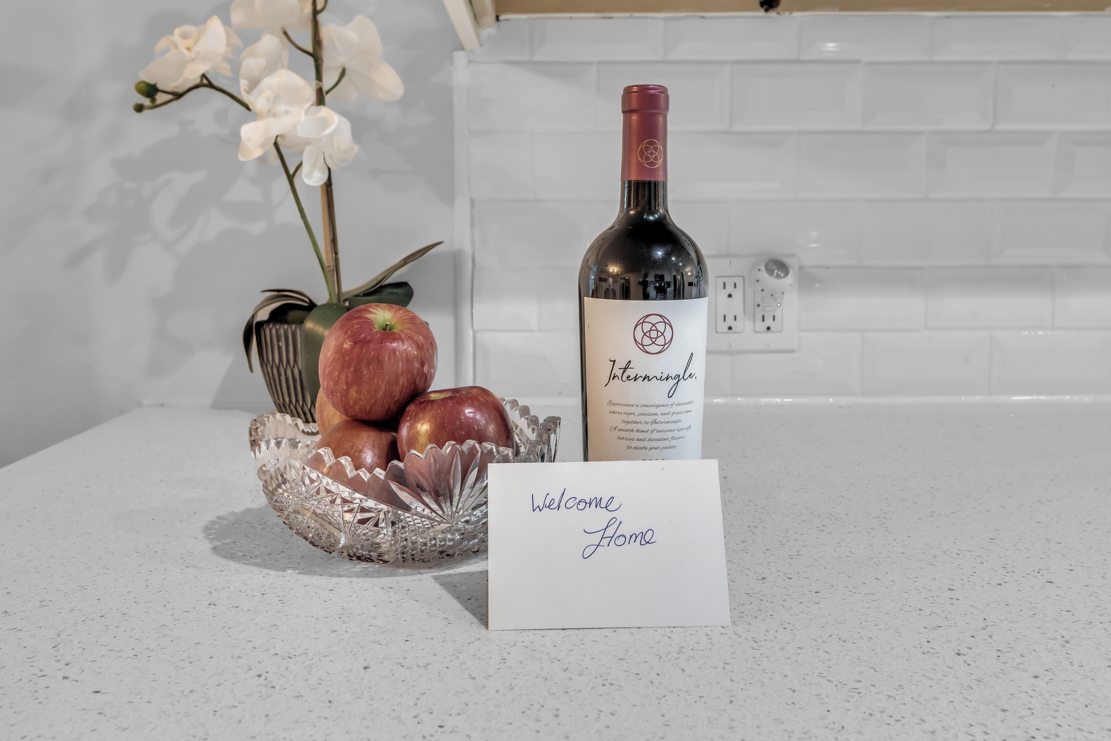 A kitchen counter with a wine bottle, a bowl of fruit, and a 'Welcome Home' card.