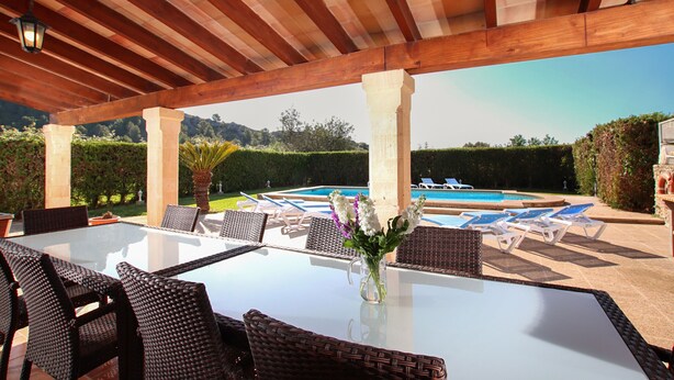 An outdoor patio with a glass table, wicker chairs, a vase with flowers, and a pool in the background. - LLORENS (IVANA).  5 bedroom villa to enjoy with family and friends (Pollensa)