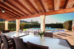 Un patio al aire libre con una mesa de vidrio, sillas de mimbre, un jarrón con flores y una piscina al fondo.