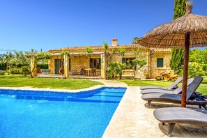 Ein Swimmingpool mit Liegestühlen, ein Steinhaus mit Ziegeldach und ein Reetdachschirm.
