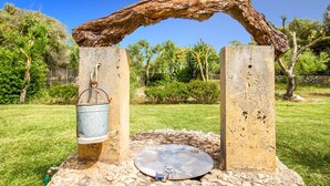 Une structure rustique en plein air, avec un seau en métal suspendu à une poutre en bois, une base en pierre et une plaque métallique posée sur le sol.