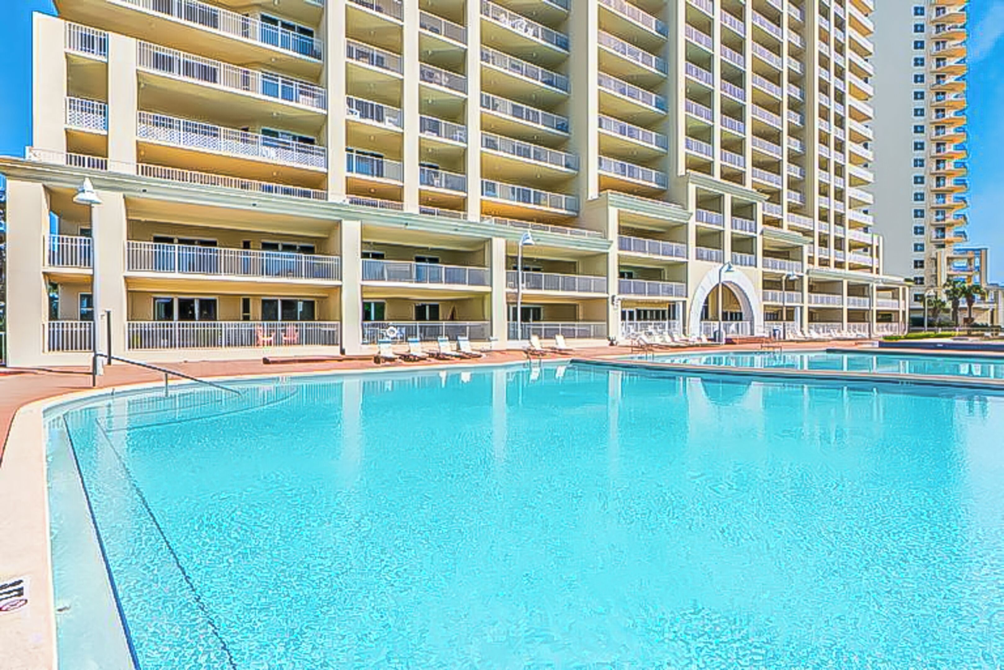 A hotel with multiple balconies, a swimming pool, and a clear sky.
