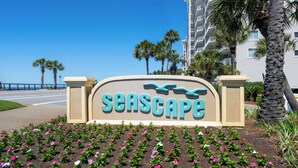 A sign with the word 'SEASCAPE' and a stylized wave design, surrounded by a garden with pink and white flowers. - Large Private Balcony ~ Spacious Living Area (Miramar Beach)