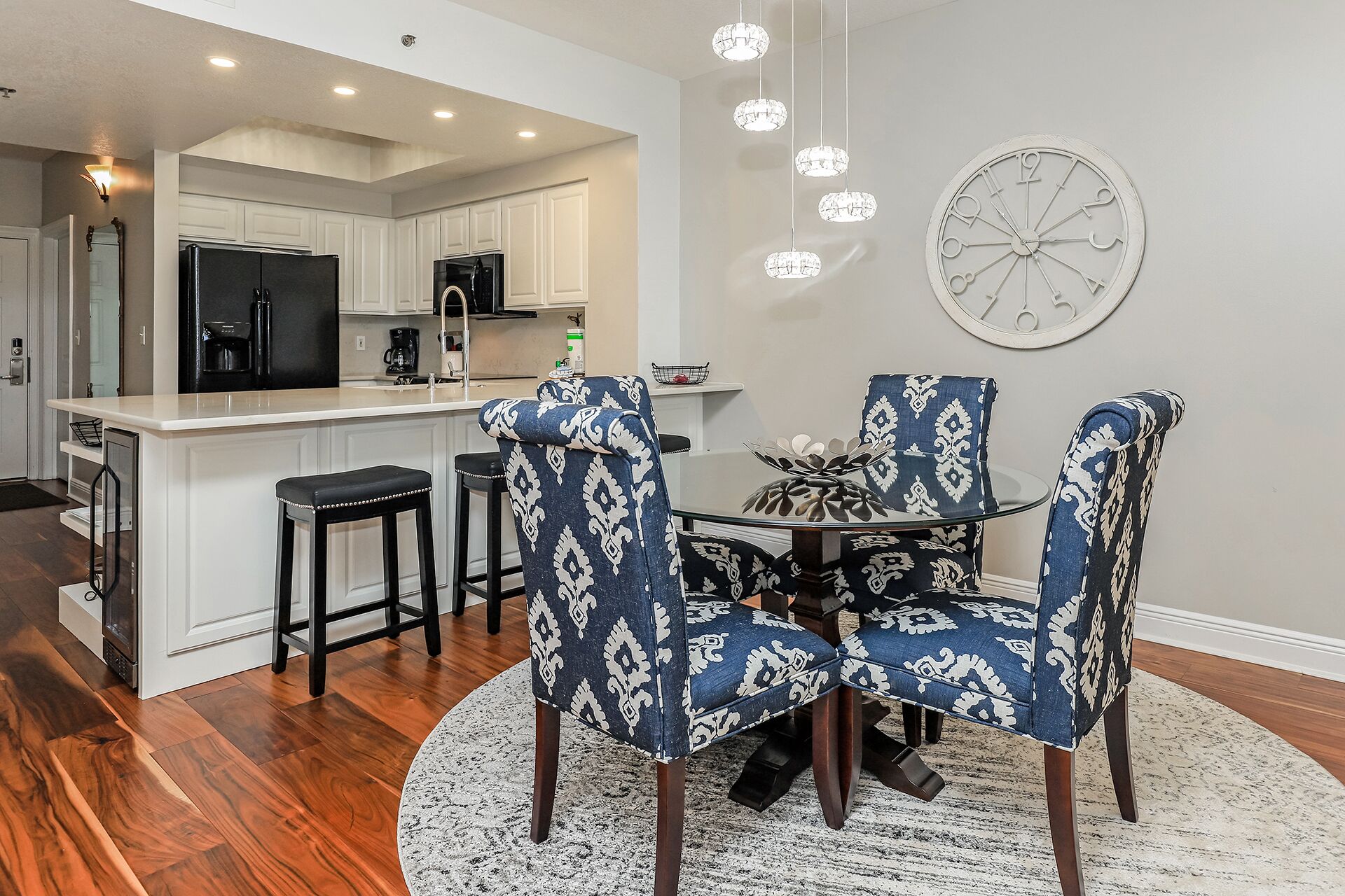 A modern kitchen with white cabinets, a black refrigerator, and a coffee maker. There is a dining area with a round table and four patterned chairs.