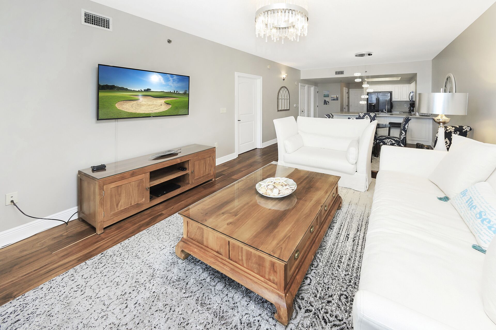 Ein modernes Wohnzimmer mit Flachbildfernseher, Couchtisch aus Holz, weißes Sofa und einer Küche im Hintergrund.